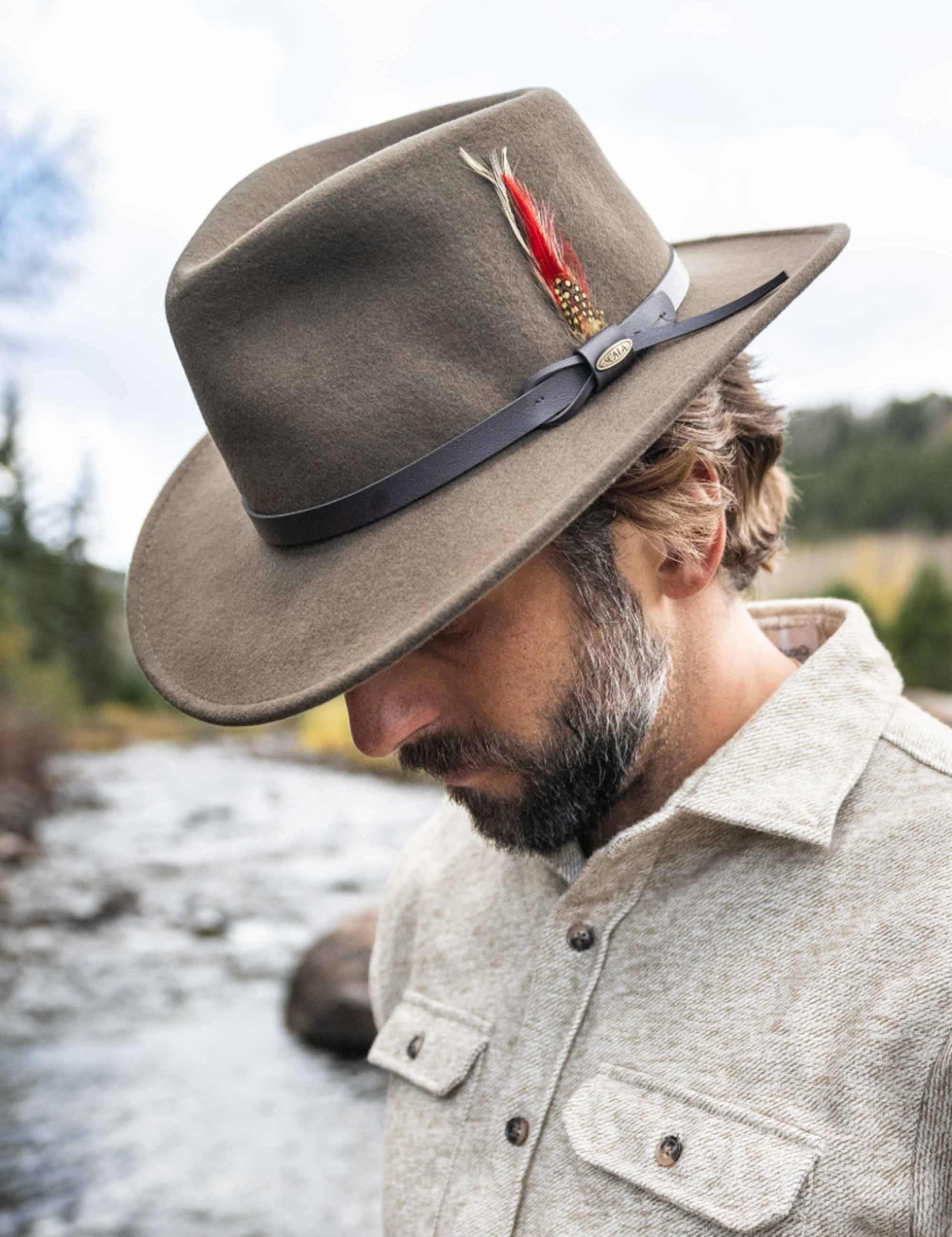 Man wearing a brown fedora hat with a feather, standing by a river.