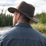 Man looking away wearing a brown long brim hat and dark blue jacket looking towards a river with trees along the way