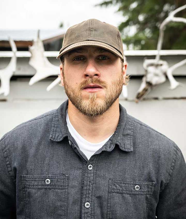 Man wearing a brown baseball cap with a white and grey shirt against a white wall with white antler bones along the top