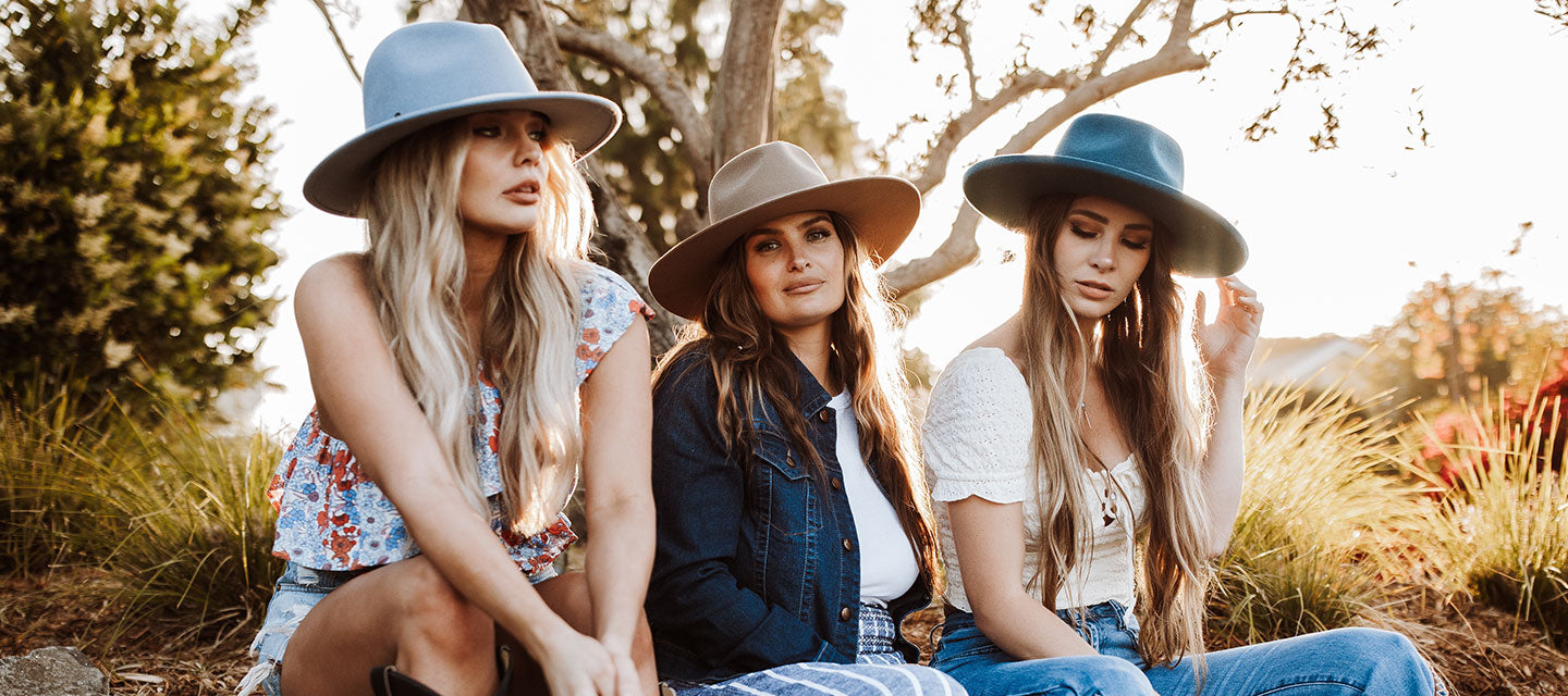 Three women wearing white and blue clothes sitting on the ground near brush and a dead tree at sunset
