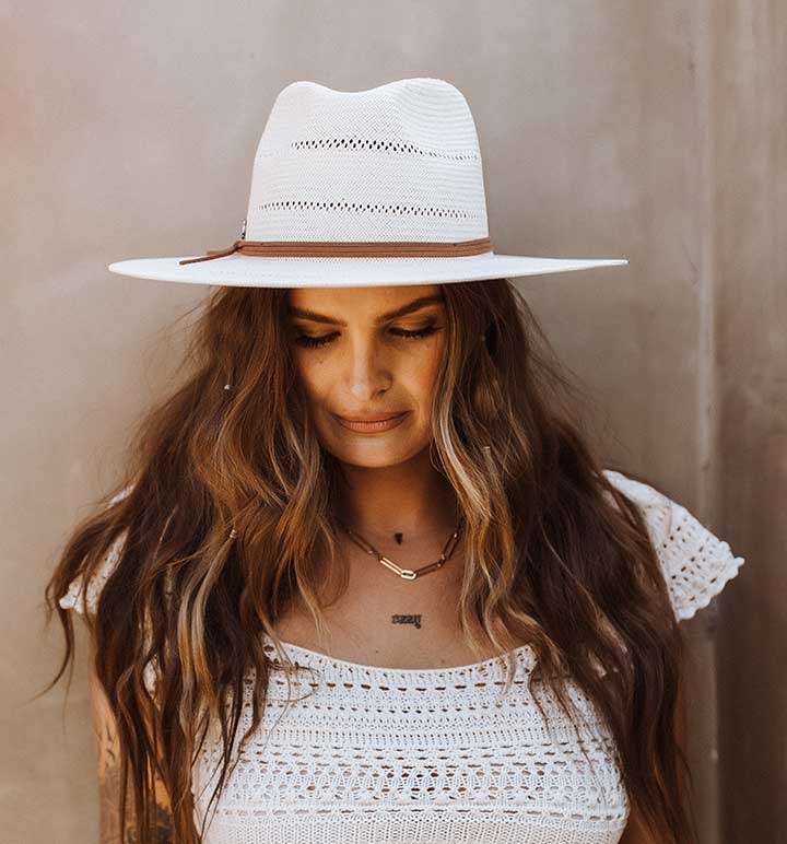 Woman wearing a white hat with brown tassels looking down at her white dress while standing against a grey wall