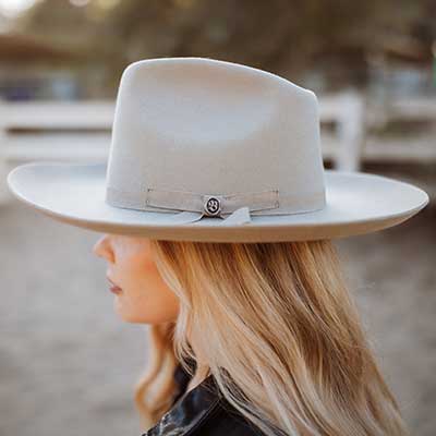 Woman wearing a grey medium brimmed hat with a small shiny Biltmore pin attached to the side on top of a ribbon