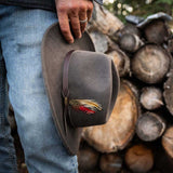 Person holding a gray fedora hat with a feather near their blue jeans against a background of stacked logs