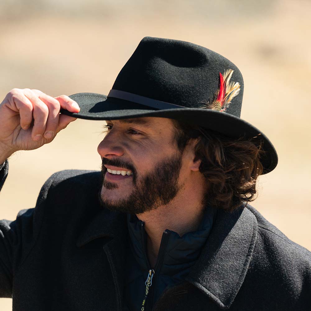 Man wearing a black hat with a feather smiling outdoors while slightly tipping his hat with his right hand