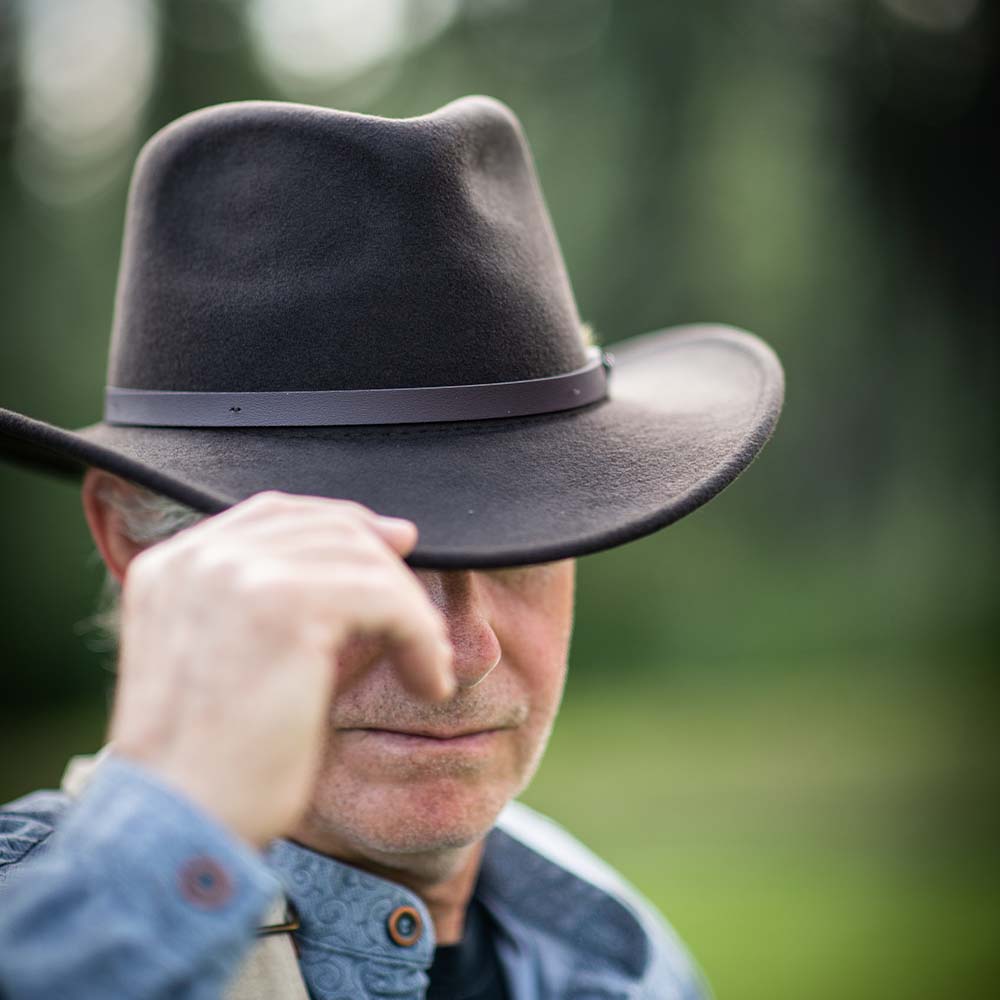 Man wearing a chocolate colored hat  while tipping it slightly downward against a blurred green background