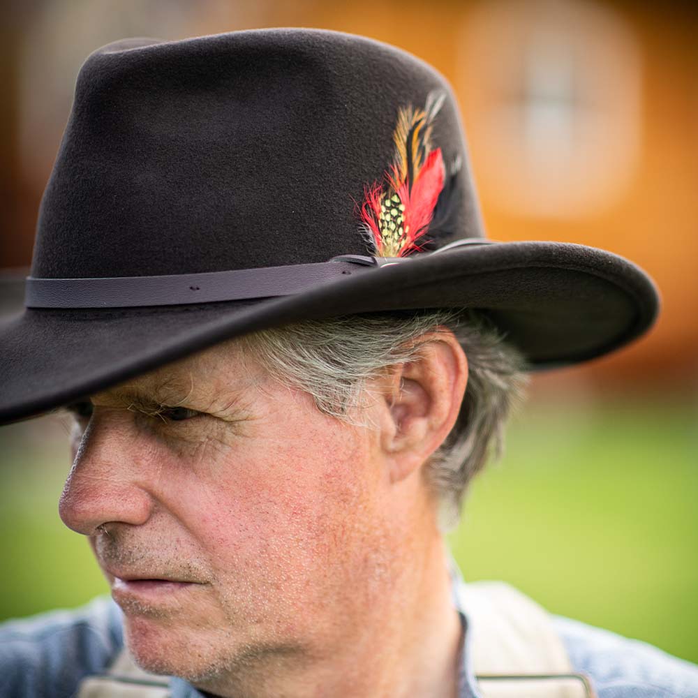 Man wearing a chocolate colored fedora hat with a feather, looking to the side with a blurred natural background