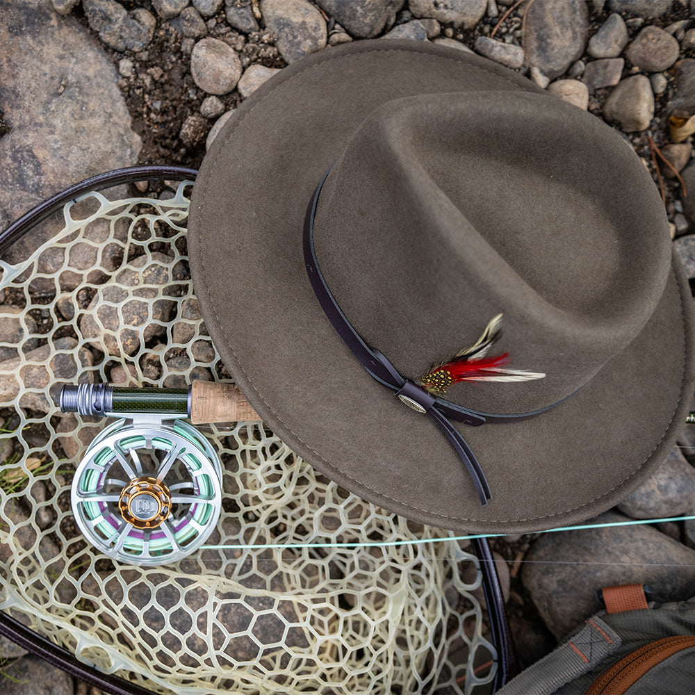 Khaki colored fedora hat with feather on a fishing net and rod on a stone surface