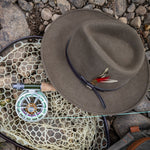 Khaki colored fedora hat with feather on a fishing net and rod on a stone surface