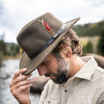 Man wearing a khaki colored fedora hat with a feather ribbon tipping his head and hat downwards against a blurred creek