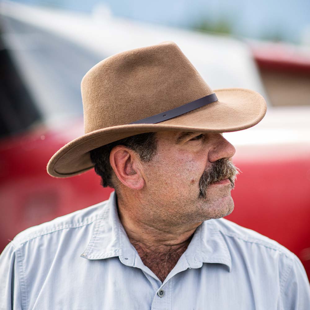 Man wearing a pecan colored fedora hat and light blue shirt with a red blurred background