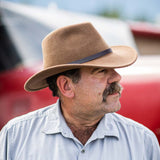 Man wearing a pecan colored fedora hat and light blue shirt with a red blurred background
