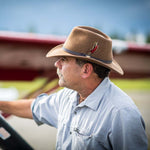 Man wearing a pecan fedora hat with a feather standing near a blurred red airplane wing
