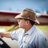 Man wearing a pecan fedora hat with a feather standing near a blurred red airplane wing
