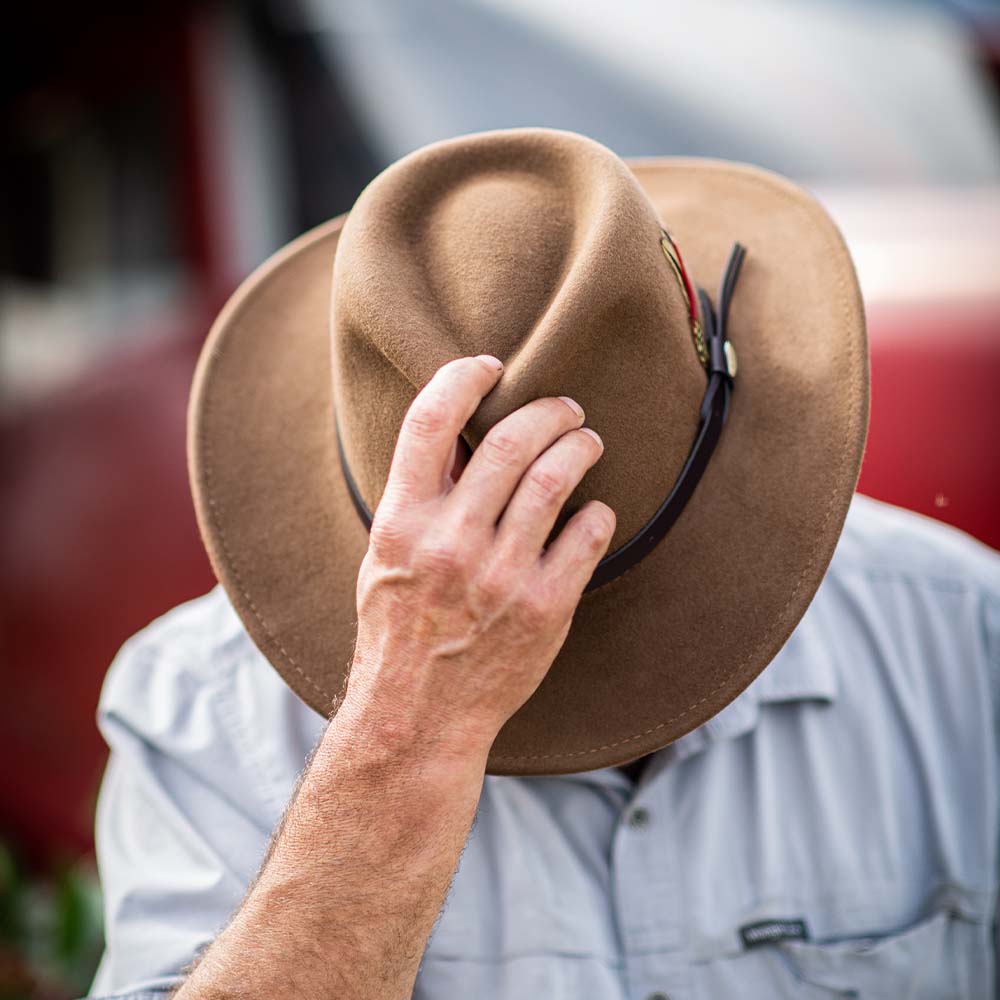 Person wearing a pecan colored hat tipping it down to cover their face against a red blurred background