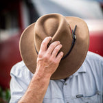 Person wearing a pecan colored hat tipping it down to cover their face against a red blurred background