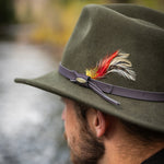 Pine colored fedora hat with feather and light colored band against a blurred natural background