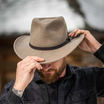 Bearded man adjusting a putty colored fedora hat against a blurred natural background