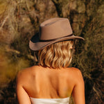 Blonde short haired woman wearing a putty colored hat in a natural setting facing away with her back
