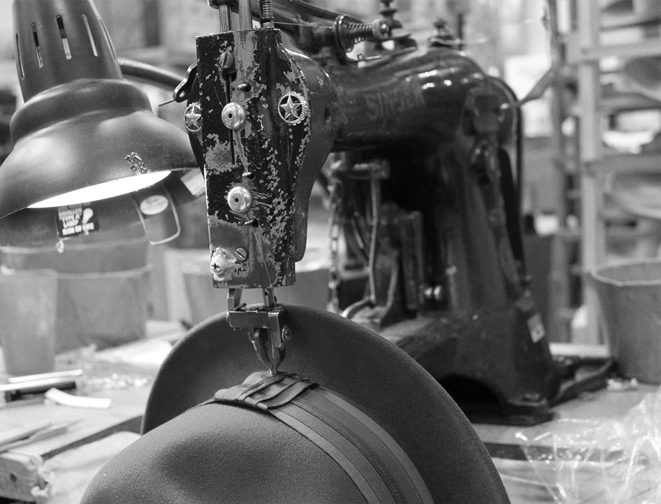 A sewing machine working on a hat band for a hat with a harsh lightbulb hovering over them