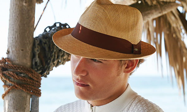 Man wearing a dark tan hat and brown band near the ocean leaning against a wooden structure