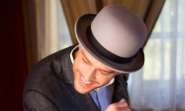 Grinning man wearing a grey bowler hat and dark jacket inside of a house near curtains and a window