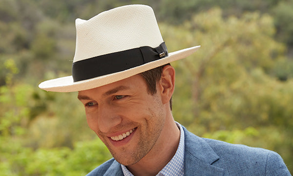 Smiling man wearing a light colored hat with black band and a blue jacket against green bushes