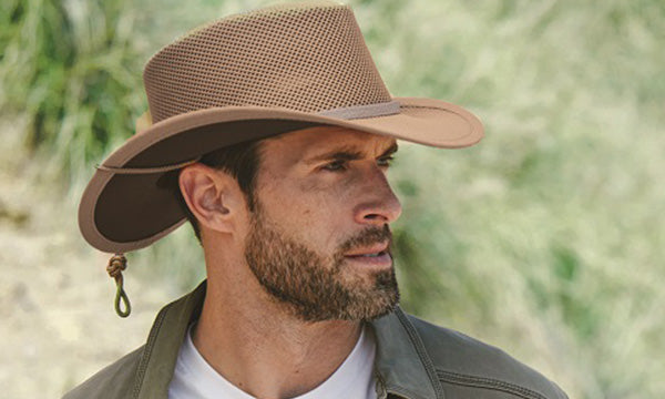 Man wearing a floppy brown hat and green jacket near a bushy trail