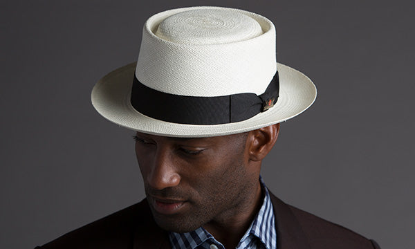 Man wearing a white hat with black band and dark colored jacket against a gray backdrop