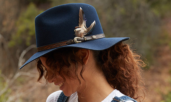 Smiling woman in a navy blue hat with a big feather against a forested backdrop