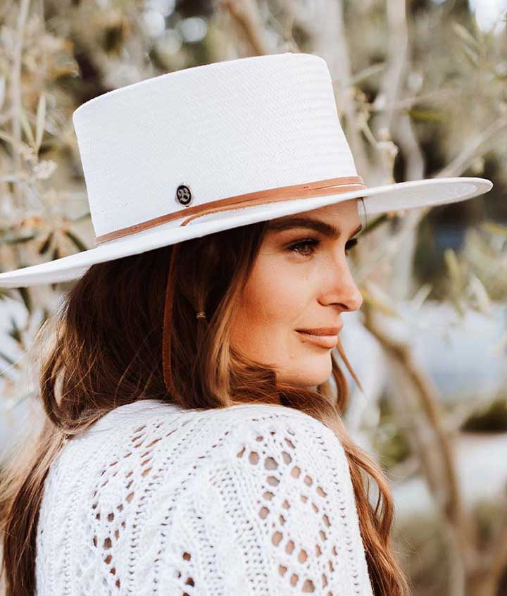 Woman wearing a white hat with light brown band and white sweater with small holes smiling against an old small tree